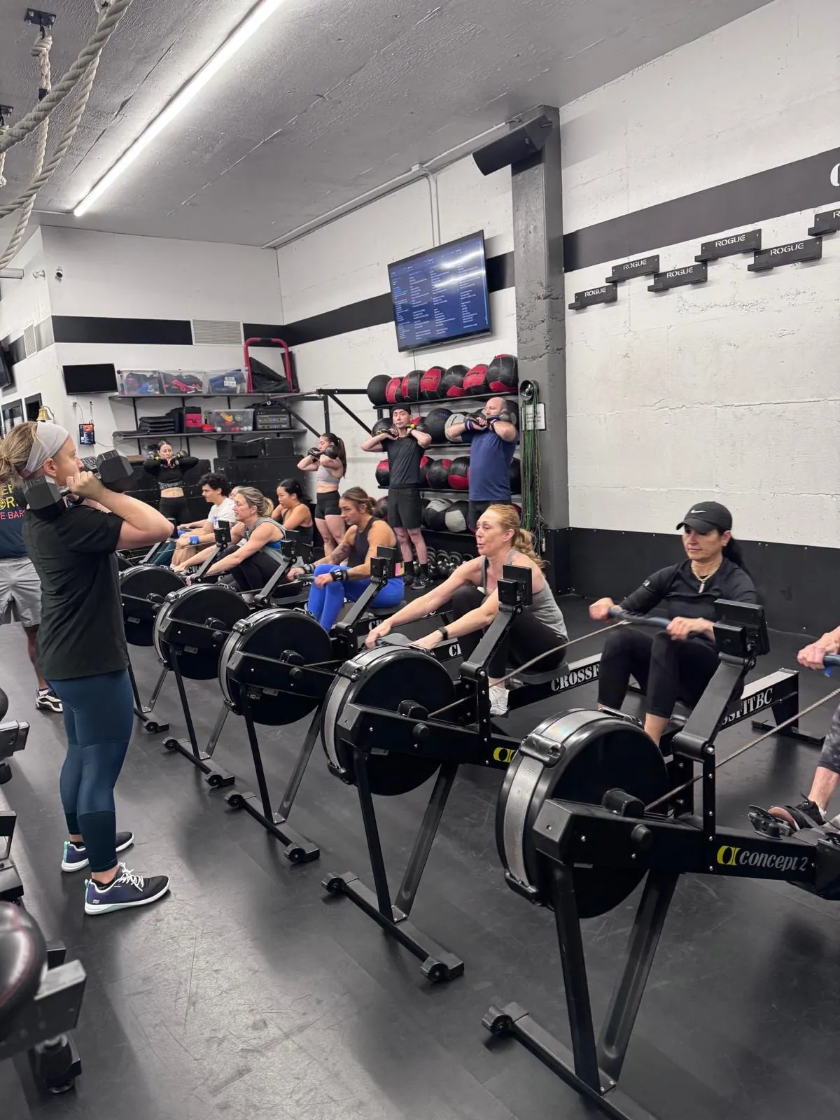 CrossFit BC members rowing in the foreground while others train with dumbbells behind them.
